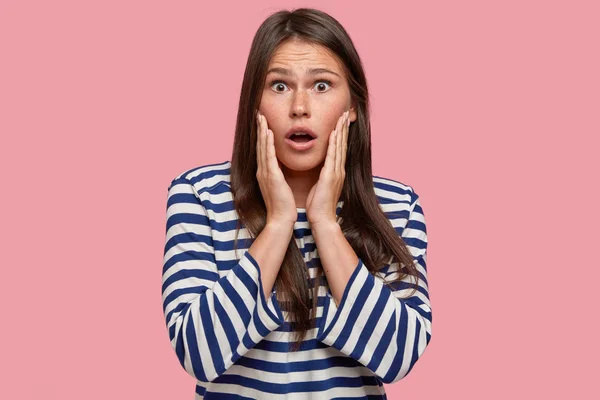 Photo of surprised female volunteer shocked with unexpected suggestions and ideas, hears amazing details of something, looks emotionally, dressed in striped sailor clothes, isolated over pink wall