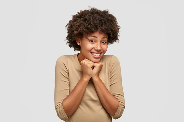 Positive black girlfriend receives good comments in her blog, keeps both hands under chin, feels pleasure, isolated over white background. African American woman rejoices pleasant moment in life