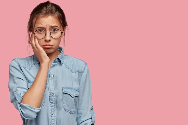 Waist up shot of beautiful dejected Caucasian female has miserable facial expression, wears stylish denim shirt, keeps hand on cheek, being in low spirit, isolated over pink background with free space