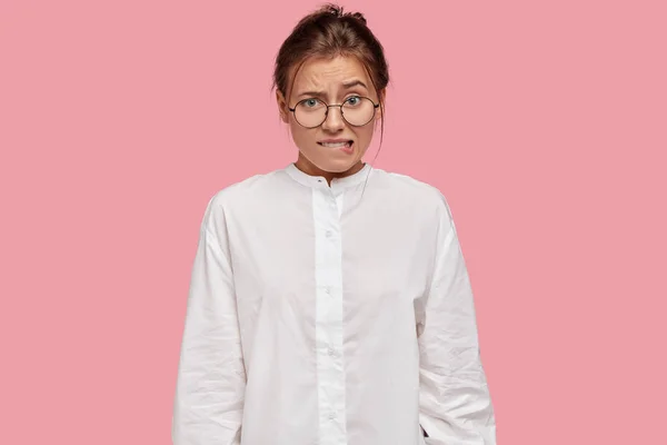 Doubtful nervous woman bites lips with embarrassed expression, raises eyebrows, has to make serious decision, wears casual white shirt, spectacles, models against pink background.