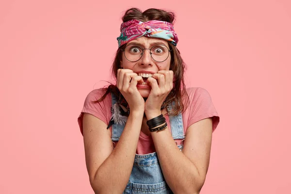 Photo of nervous anxious hippy woman bites finger nails, wears headband, round spectacles, dressed in denim overalls, poses over pink background, being frustrated.