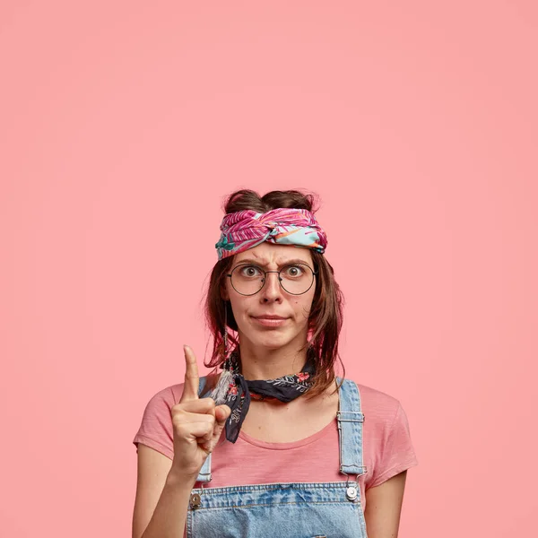 Photo of serious angry hippie being discontent with noisy neighbors, points with index finger upwards, wears stylish headband and denim overalls, free space against pink background for your text