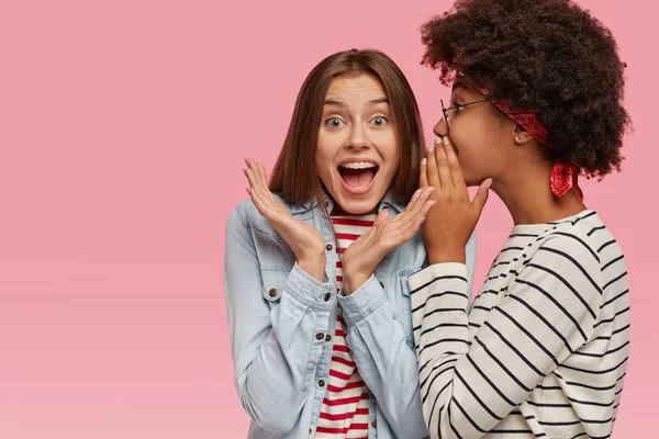 Emotional European woman clasps hands and exclaims loudly as hears rumors from best friend. Afro American woman whispers secret at companions ear, stands sideways, gossip together, stand indoor