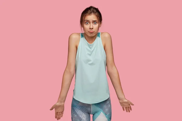 Horizontal shot of puzzled young European woman shrugs shoulders, purses lower lip, wears sportswear, poses against pink studio background.