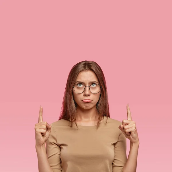 Photo of impressed female model feels unhappy, keeps both hands up, shows with index fingers something above head, purses lips, displeased seeing some item, isolated over pink studio background