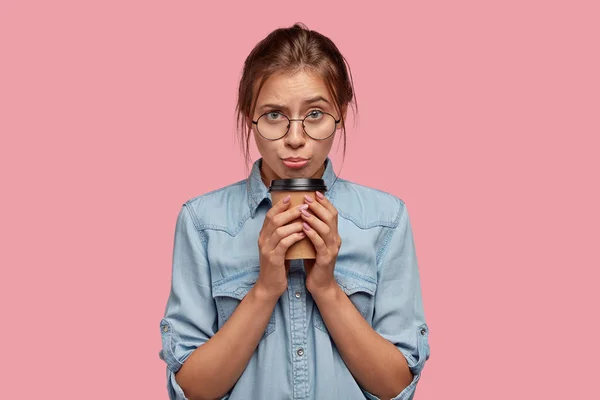 Discontent Caucasian young woman frowns face, has displeased facial expression, holds takeaway coffee in both hands, sad to have much work, dressed in denim shirt, isolated over pink background