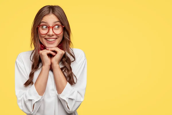 Positive Caucasian woman with tender smile, keeps hands under chin, giggles positively aside, dressed in white shirt, stands against yellow background with free space for your promotional content