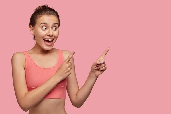 Horizontal shot of pretty young woman with perfect body, indicates with both index fingers aside, being glad to show some item, isolated over pink background.