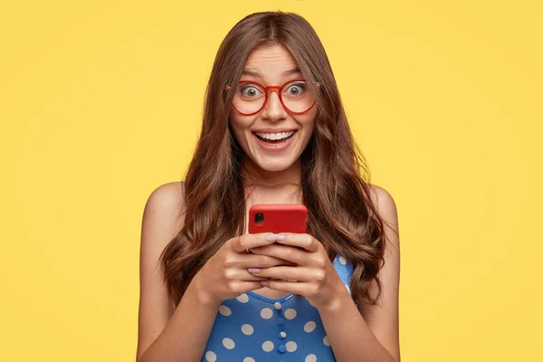 Shot of cheerful female model wears glasses, holds cell phone, takes pictures, talks via video message, has nice friendly talk, dressed in fashionable outfit, isolated over yellow background