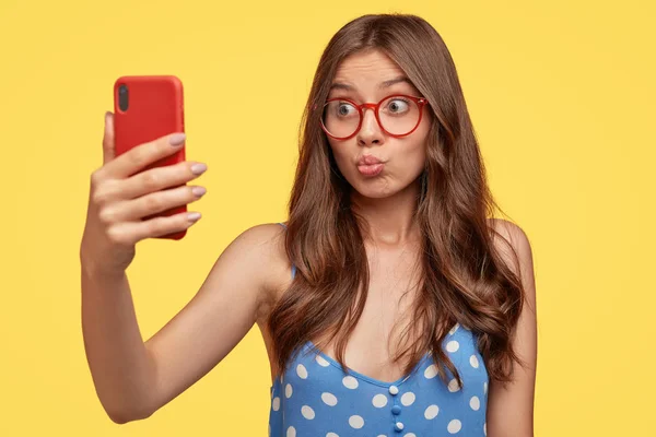 Pretty girl takes selfie, pouts lips at camera of cell phone, makes video call, shoots something for blog, takes photo of herself, wears glasses.