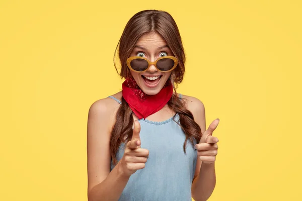 I have nice plans on you. Joyful fashionable girl points with both index fingers directly at camera, hints on something wonderful, wears trendy sunglasses, isolated over yellow studio background.