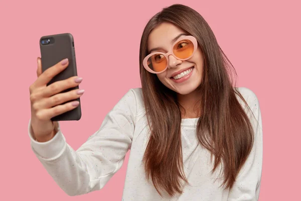 Horizontal shot of beautiful dark haired girl smiles happily, tilts head, holds cell phone, records vlog, takes selfie, wears trendy sunglasses and white casual sweater, isolated over pink background