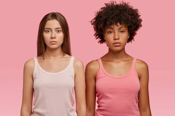 Studio shot of multiracial women in casual vest, have serious look at camera, stand next to each other against pink background, going to have sport training together.