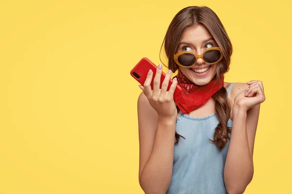 Pleased delightful female wears trendy shades, holds mobile phone, listens pleasant melody, enjoys high speed internet, waits for important call, isolated over yellow background with copy space