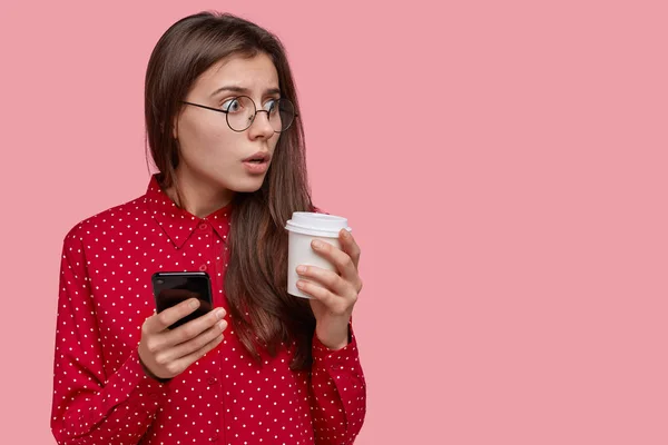 Indoor shot of puzzled surprised dark haired beautiful woman holds take out coffee, modern cell phone, dressed in red blouse, astonished to notice something aside, stands sideways over pink background