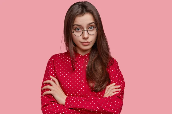 Self confident serious young lady keeps arms folded, dressed in polka dot outfit, looks attentively directly at camera, wears round glasses, listens information from interlocutor, isolated on pink