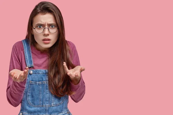 Indignant Caucasian woman looks angrily at camera, gestures with both hands, wears spectacles, looks confused and shocked, blank space for your promotional content