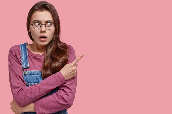 Questioned woman with surprised expression points aside with index finger, opens mouth from amazement, watches amazing show, feels impressed, isolated over pink background, can not say word.