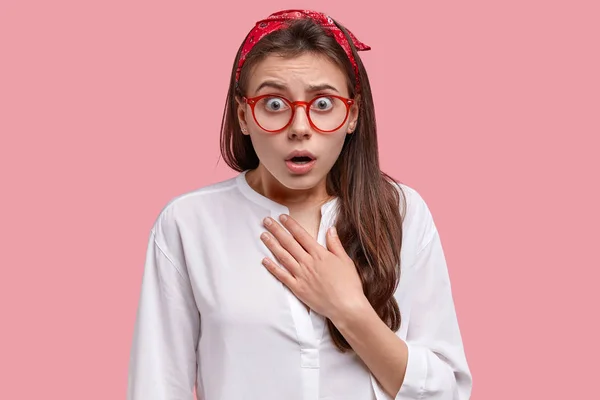 Studio shot of emotional woman gazes in stupor, afraid of something terrible, dressed in white oversize shirt and headband, models over pink background.