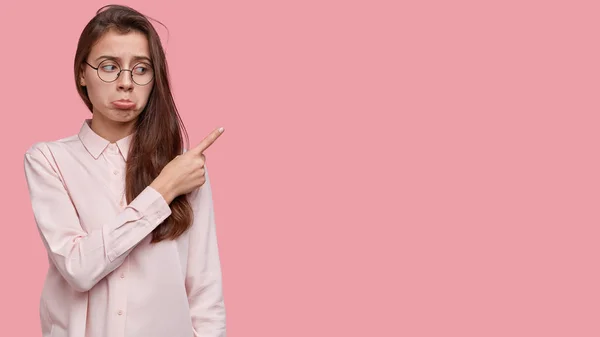 Unimpressed dejected woman purses lips, points aside upwards, being dejected, wears transparent glasses and shirt, stands over pink studio wall with blank space for your advertising content.