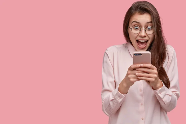 Studio shot of pretty woman stares at mobile phone, feels satisfied, stares at screen, opens mouth, dressed in formal shirt, isolated over pink background with blank space for your promotion