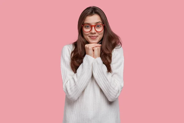 Photo of delighted pretty young woman with long hair, keeps hands under chin, wears red rim spectacles and white long jumper, listens delightfully, isolated over pink background.