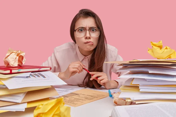 Puzzled dark haired lady purses lips, surrounded by heaps of documents, attends refresher course, wears optical glasses, isolated over pink background, works indoors.