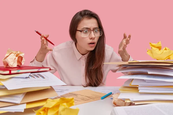 Frustrated brunette lady raises hands with bewilderment and shock, stares at heap of papers, doesnt know from what to start working, holds pen, isolated over pink studio wall prepares financial report
