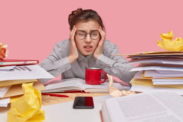 Close up shot of dissatisfied woman keeps hands on temples, frowns face, has fatigue expression, wears round spectacles, prepares statistics report, isolated over pink background.