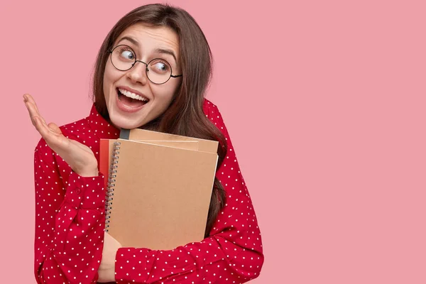 Happy indecisive woman looks aside with clueless expression, holds spiral notepad, cant decide on day plans, thinks about starting freelance career, isolated over pink background with copy space