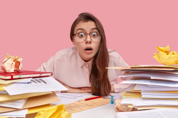 Horizontal view of surprised dark haired emotive female analyst has surprised gaze at camera, wears big spectacles, dressed in shirt, surrounded with paper documents, isolated over pink background