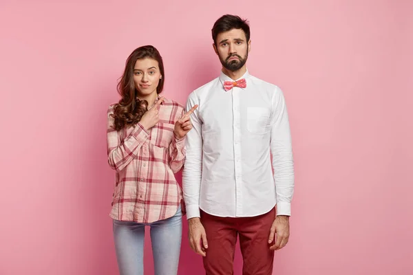 Photo of dissatisfied woman and man stand closely, discontent with new outfit of guy, isolated over pink background. Pretty dark haired lady points at bearded male companion in elegant clothes