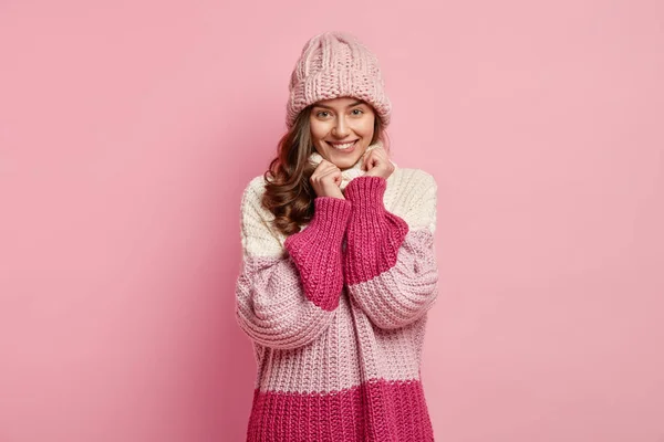 Photo of happy delighted young woman with gentle smile, dressed in woolen sweater, hat, keeps hands under chin, isolated over pink background, looks with intriguing expression.