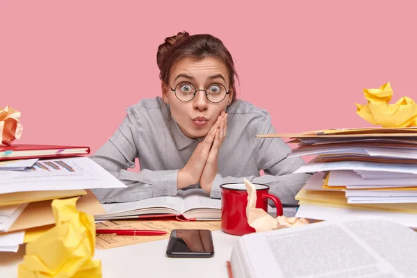 Indoor shot of attractive student keeps eyes widely opened, lips folded, wears optical glasses, dressed in stylish shirt, sits at desktop with many papers and textbooks, drinks coffee, uses cellular
