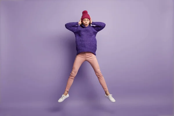 Horizontal shot of surprised terrified woman stares with bugged eyes, feels overwhelmed, jumps high in air, wears warm winter hat, knitted jumper and white sport shoes, poses over purple wall