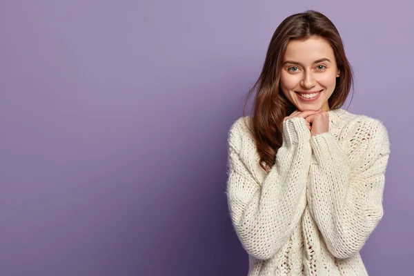 Pleased beautiful lady with toothy gentle smile, has healthy skin, green eyes, long straight hair, holds hands under chin, wears warm white jumper, happy to be photographed, stands over lilac wall