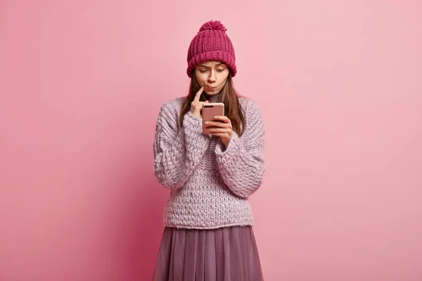 Attentive focused woman purses lips, focused at smart phone, reads something with puzzled ending, connected to wireless internet, dressed in stylish clothes for winter, isolated over pink background