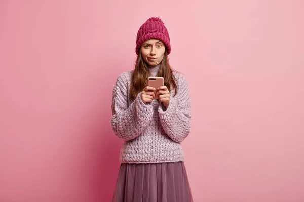 Upset dissatisfied young woman purses lips, holds modern cell phone, sends text messages in social networks, wears warm headgear, loose winter sweater and skirt, poses against rosy background