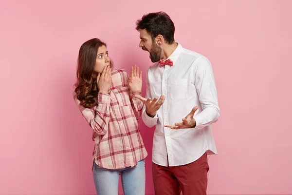 Scared guilty woman stares at angry boyfriend who shouts at her, suspects in betrayal, gestures with irritation, dressed in elegant shirt with bowtie isolated over pink background. What have you done?
