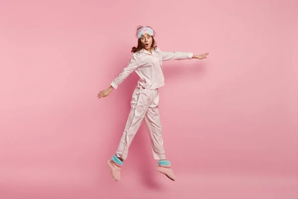 People, morning and amazement concept. Frightened Caucasian woman with shocked expression, wears pyjamas, jumps high, spreads hands, isolated over pink background, stunned by horrified thing