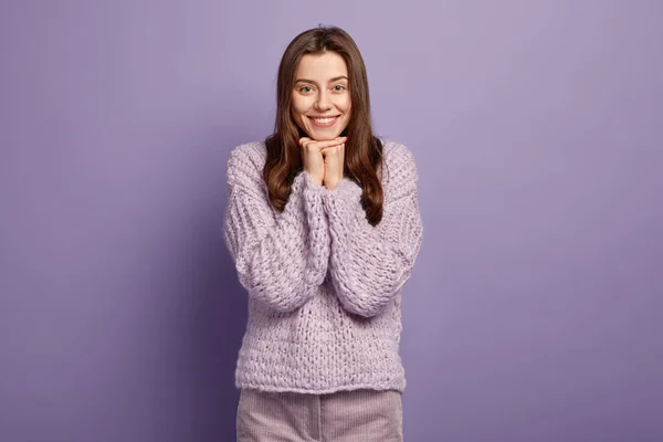 Horizontal shot of tender pleased European lady keeps hands together, smiles happily, shows white teeth, dressed in loose sweater in one tone with background, isolated over purple background