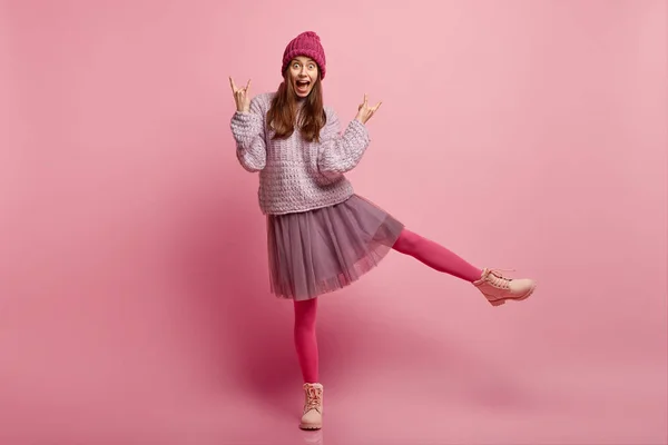 Fashionable cool woman with joyful expression, dressed in hat, sweater and skirt, shows rock n roll gesture, raises legs, poses over pink background with free space for your advertisement or text