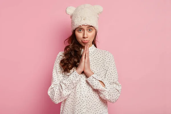 Horizontal shot of pleading attractive woman with green eyes, keeps palms pressed together, has pity look, asks for apology, wears funny hat in form of bear, polka dot shirt, isolated on pink wall