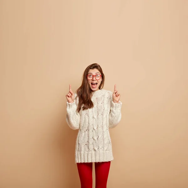 Glad optimistic young woman with joyful expression, shows direction upwards, points with fore fingers, wears long sweater and red leggings, isolated over beige studio, with mock up place for your text