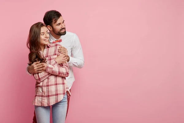 Horizontal shot of positive European couple in love embrace and focused aside, notice funny scene, enjoy togetherness, pose against pink background with blank space for your promotional content.