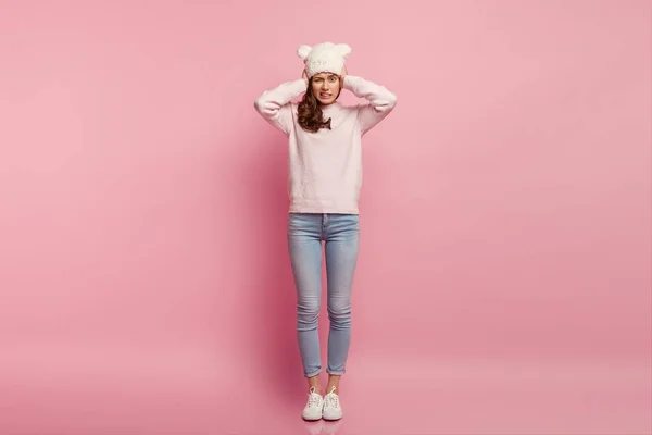 I don\'t like this sound. Frustrated displeased woman covers ears, feels dissatisfied and annoyed with loud noise, wears funny white hat with ears, jumper, jeans and sportshoes, isolated on pink wall