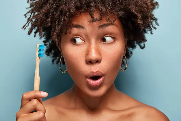 Healthy wondered woman shows wooden toothbrush, has curly hairstyle, keeps mouth opened, does everyday morning routine, daily oral care, surprised to have problems with teeth.