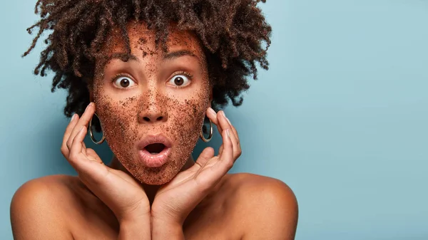 Image of stupefied woman astonished to find out about skin problems, removes pores with coffee scrub, has eyes popped out, touches cheeks, models topless, isolated on blue wall.