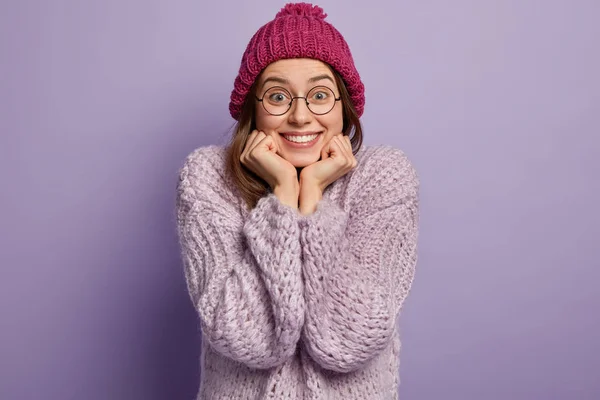 Indoor shot of happy smiling woman keeps hands under chin, shows white teeth, enjoys winter weather, going to have walk, wears hat with pompon, oversized sweater, being in good mood, isolated on lilac