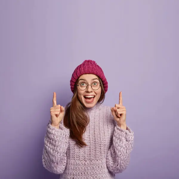 Check out this cool place. Optimistic European woman with joyful gleeful expression points up using fore fingers, wears winter hat and jumper, notices something amazing on ceiling gazes at nice sunset